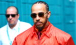 Man with braided hair and a goatee wearing a bright red windbreaker, sunglasses, and a maroon tie; blurred figure in background.