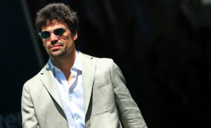 Man wearing a light gray blazer and white shirt, sunglasses, outdoors on a sunny day.