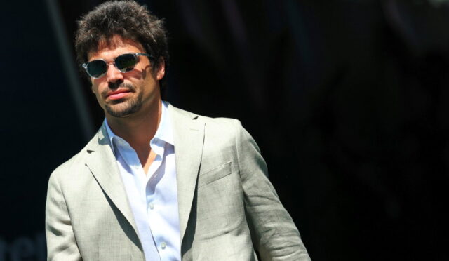 Man wearing a light gray blazer and white shirt, sunglasses, outdoors on a sunny day.
