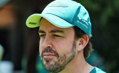 Close-up of a man with a teal cap and beard looking to the left, outdoors at a sunny event.