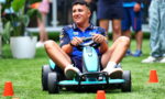 Young man in a blue shirt drives a turquoise go-kart through orange cones on a grassy field, smiling as he participates in an outdoor event.