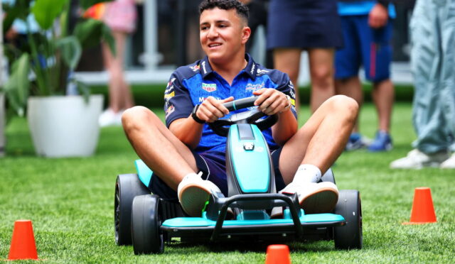 Young man in a blue shirt drives a turquoise go-kart through orange cones on a grassy field, smiling as he participates in an outdoor event.