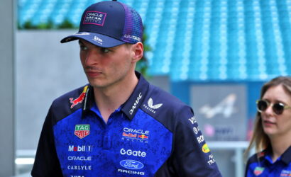 Professional race driver in Oracle Red Bull Racing gear, blue polo and cap, standing in a pit/track area and looking to the left.