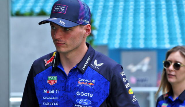 Professional race driver in Oracle Red Bull Racing gear, blue polo and cap, standing in a pit/track area and looking to the left.