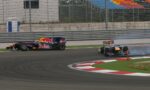 Two Formula 1 cars race through a corner, a Red Bull car leading while the blue-red car hugs the inside with tire smoke in the background.