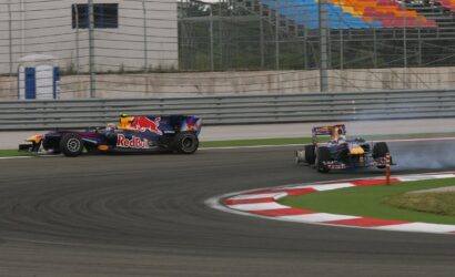 Two Formula 1 cars race through a corner, a Red Bull car leading while the blue-red car hugs the inside with tire smoke in the background.