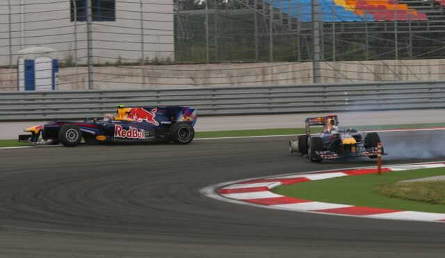 Two Formula 1 cars race through a corner, a Red Bull car leading while the blue-red car hugs the inside with tire smoke in the background.