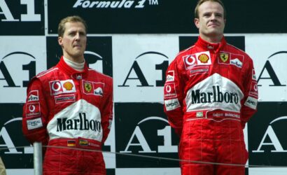 Two Ferrari Formula 1 drivers in red Marlboro racing suits stand on a podium, facing forward against a black-and-white backdrop with sponsor logos.