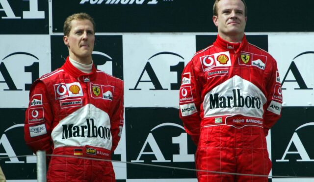 Two Ferrari Formula 1 drivers in red Marlboro racing suits stand on a podium, facing forward against a black-and-white backdrop with sponsor logos.