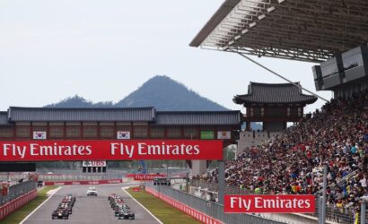 Race cars line up on a track as crowds watch from grandstands, with bold Fly Emirates banners overhead.