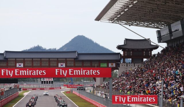 Race cars line up on a track as crowds watch from grandstands, with bold Fly Emirates banners overhead.