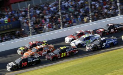 A pack of stock cars racing on a track with a crowded grandstand in the background, car numbers 78, 19, 24, 13, and 5 visible in the lead.