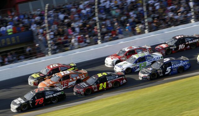 A pack of stock cars racing on a track with a crowded grandstand in the background, car numbers 78, 19, 24, 13, and 5 visible in the lead.