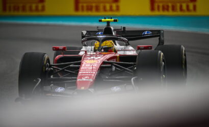 Red Formula 1 car on a race track with a yellow-helmeted driver, Pirelli banners in the background