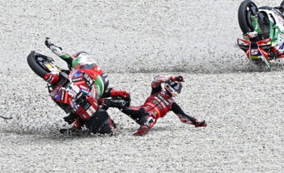 Two MotoGP riders crash on a gravel track, one tumbling headfirst while another bike skids nearby in full racing gear.
