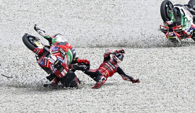 Two MotoGP riders crash on a gravel track, one tumbling headfirst while another bike skids nearby in full racing gear.
