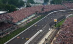 Wide-angle view of a crowded motorsport race with cars on a curved track and packed grandstands on both sides.