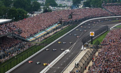 Wide-angle view of a crowded motorsport race with cars on a curved track and packed grandstands on both sides.