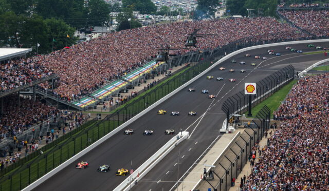 Wide-angle view of a crowded motorsport race with cars on a curved track and packed grandstands on both sides.