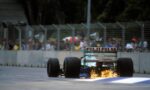 Rear-view of a Formula 1-style race car with sparks shooting from underneath as it speeds along the track; crowd behind a safety fence watches.