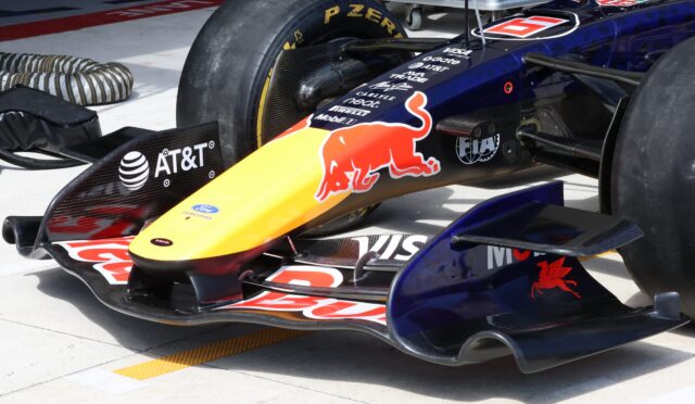 Close-up of a Red Bull Formula 1 car nose in the pit area, showing sponsor decals and the front wing authority plate area on a yellow-to-red livery.