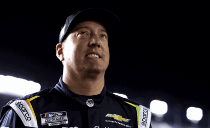 Male race car driver in a dark NASCAR suit with Chevrolet logo, looking upward at bright stadium lights at night night race backdrop.