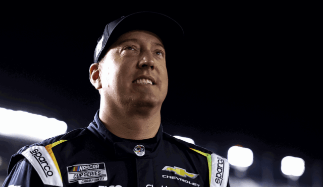 Male race car driver in a dark NASCAR suit with Chevrolet logo, looking upward at bright stadium lights at night night race backdrop.