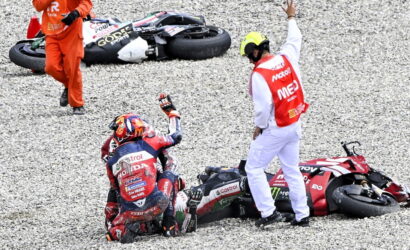 Motocycle racer sits on gravel after a crash while a marshal in a red-and-white vest raises a hand nearby and fallen bikes lie beside them on the track surface.
