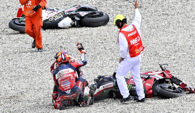 Motocycle racer sits on gravel after a crash while a marshal in a red-and-white vest raises a hand nearby and fallen bikes lie beside them on the track surface.