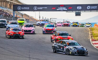 Race cars on a curving track preparing to start a race under a banner reading Turkish Grand Prix 2021/2023, with spectators in stands on the left and a gray Audi leading on the front-right corner.