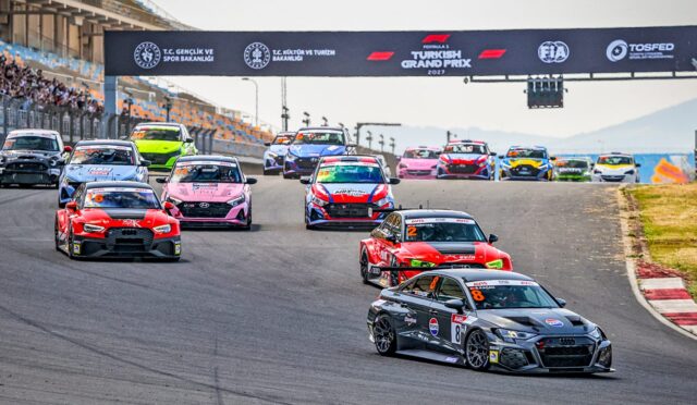 Race cars on a curving track preparing to start a race under a banner reading Turkish Grand Prix 2021/2023, with spectators in stands on the left and a gray Audi leading on the front-right corner.