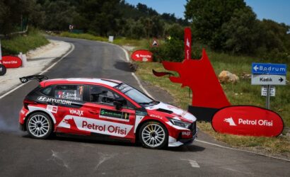 Rally car in red and white livery turning on a rural winding road with Petrol Ofisi branding and sponsor banners nearby.