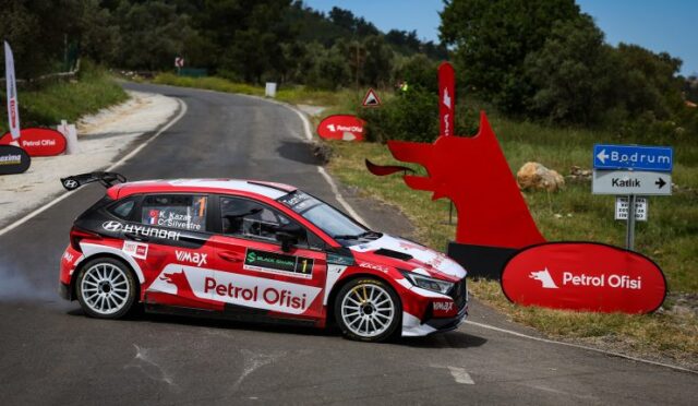 Rally car in red and white livery turning on a rural winding road with Petrol Ofisi branding and sponsor banners nearby.