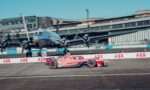 Pink Porsche race car #51 speeds along a track with a large helicopter and airport-style buildings in the background.