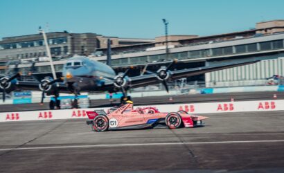 Pink Porsche race car #51 speeds along a track with a large helicopter and airport-style buildings in the background.