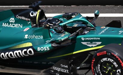 Close-up of a teal Formula E race car cockpit with a driver in a helmet, showing sponsor logos like Coherent(codec?) Cognizant, BOSS, Valvoline, and Aston Martin on the car body and wheel.