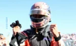 Race car driver in helmet and racing suit, fists raised in celebration on a track with spectators in the stands behind.
