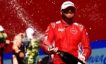 Race car driver in a red suit sprays champagne on the podium, with a blurred crowd in the background.