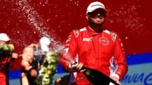 Race car driver in a red suit sprays champagne on the podium, with a blurred crowd in the background.
