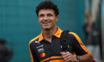Young man in an orange-and-black polo with sponsor logos, carrying a backpack and smiling at the camera.