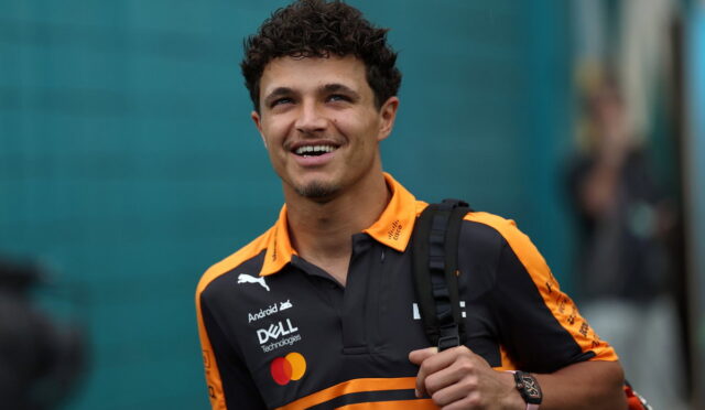 Young man in an orange-and-black polo with sponsor logos, carrying a backpack and smiling at the camera.