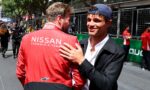 Two men share a hug at a racing event, one wearing a red Nissan Formula team jacket, seen from behind.