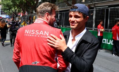 Two men share a hug at a racing event, one wearing a red Nissan Formula team jacket, seen from behind.