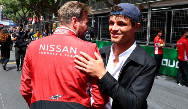 Two men share a hug at a racing event, one wearing a red Nissan Formula team jacket, seen from behind.