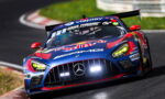 Mercedes-AMG race car in blue and red Red Bull livery turns hard into a corner on a race track, with a large rear wing and bright headlights visible.