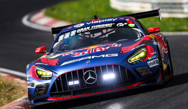 Mercedes-AMG race car in blue and red Red Bull livery turns hard into a corner on a race track, with a large rear wing and bright headlights visible.
