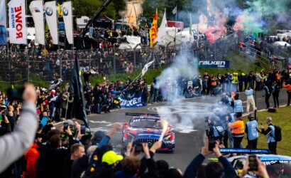 Crowd cheers along a race track as colorful smoke and racing cars speed by, with banners and tents in the background.