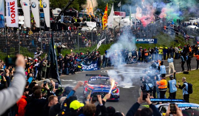 Crowd cheers along a race track as colorful smoke and racing cars speed by, with banners and tents in the background.