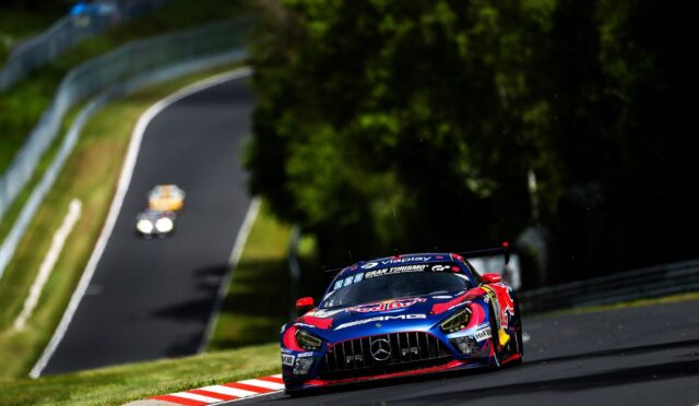 Race car speeding around a curve on a racetrack with green grass and guardrails in the background, motion blur emphasizing speed.