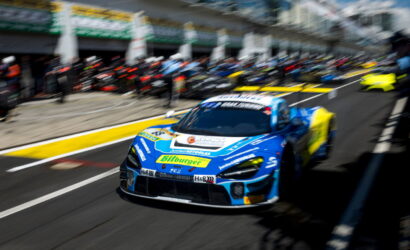 Blue race car with sponsor logos speeding from the pit lane amid a blurred crowd and garages in the background.